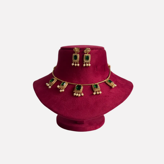 Gold-toned traditional Indian jewelry set including a necklace and matching earrings with green rectangular stones and gold bell details, displayed on a magenta velvet jewelry bust against a light gray background.