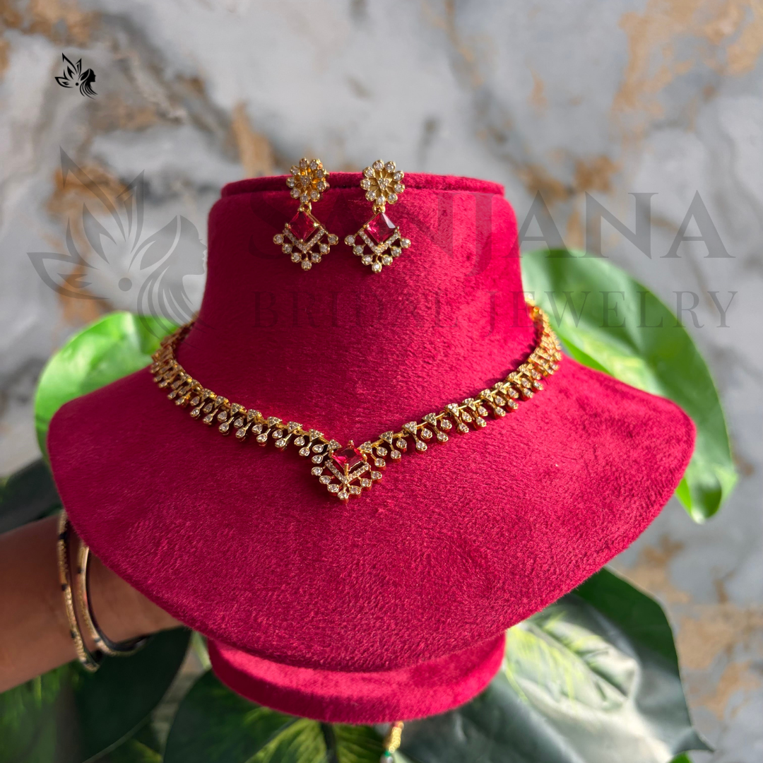 Gold-toned traditional Indian jewelry set including a necklace, earrings, and a matching stand, displayed on a bright pink velvet stand with green leaves in the background.