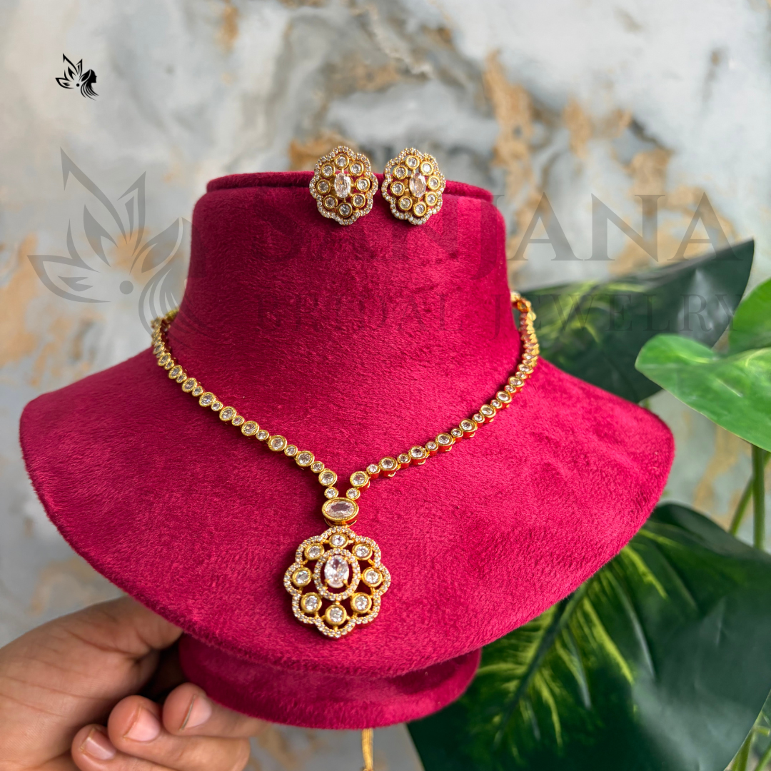 Gold-toned jewelry set with a necklace and matching earrings displayed on a magenta velvet stand, held by a hand against a marble and green leaf background.
