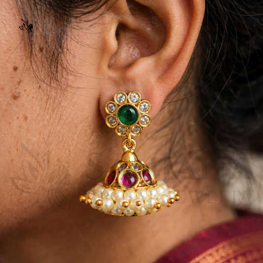 A close-up of a person's ear wearing a gold-plated traditional Indian jhumka earring with a green stone, clear stones, and white pearl-like beads.