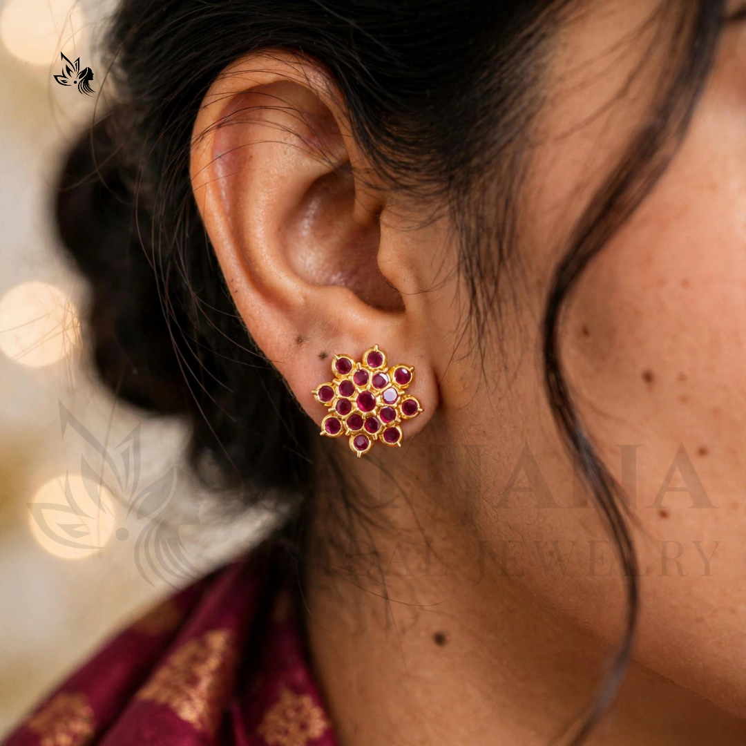 A close-up of a person's ear wearing a gold-toned floral stud earring with red stones and small clear accent stones. The background is softly blurred with warm bokeh lights, and a faint 'Anja Jewelry' watermark is visible.