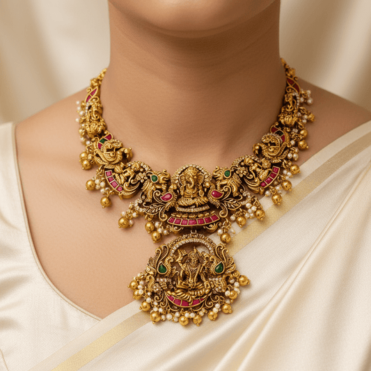 An ornate gold-toned traditional necklace with a large central pendant, decorated with green and pink enamel details and small pearl-like beads, worn by a model in a cream-colored satin garment.