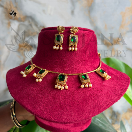 Gold-toned traditional Indian jewelry set including a necklace, earrings, and a matching hair pin, displayed on a magenta velvet stand. The jewelry features rectangular green stones and dangling pearl-like beads.