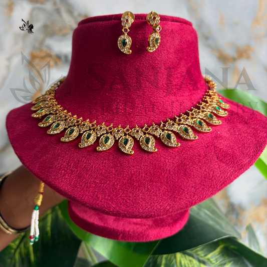 Gold-toned traditional jewelry set featuring a necklace and matching earrings with green stone accents, displayed on a magenta velvet stand. A hand is visible holding the stand, and decorative leaves and a marble-patterned backdrop are in the background.