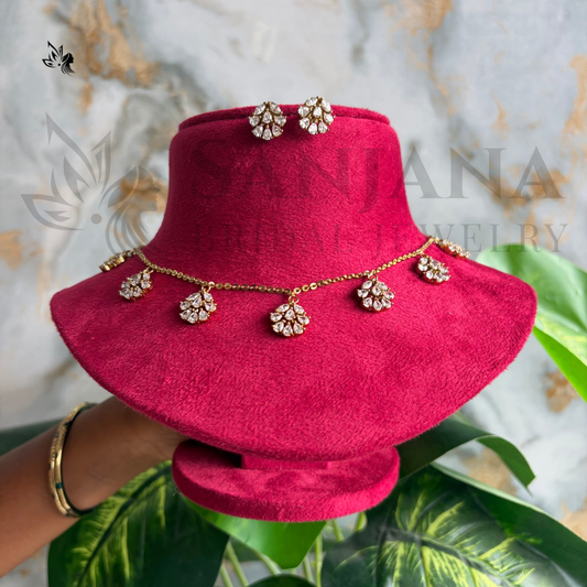 A gold-tone necklace with matching flower-shaped stud earrings displayed on a bright pink velvet jewelry bust. The necklace features five floral clusters with clear rhinestones, and a hand is holding the display stand.