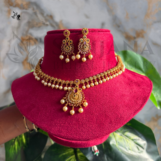 Gold-toned traditional Indian jewelry set including a necklace with a large pendant, matching earrings, and a bracelet, displayed on a bright pink velvet jewelry bust. The jewelry features ornate filigree work, gold-toned details, and small pearl-like bead drops.