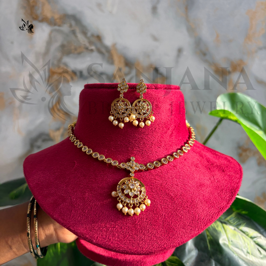Gold-toned traditional Indian jewelry set with a necklace, matching earrings, and a bracelet displayed on a bright pink velvet jewelry stand. The set features ornate paisley-style motifs with small pearl-like bead drops.