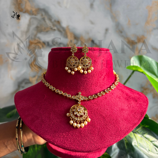 Gold-toned traditional Indian jewelry set including a necklace, matching earrings, and a bracelet displayed on a bright pink velvet stand. The jewelry features ornate filigree details and small pearl-like bead drops.