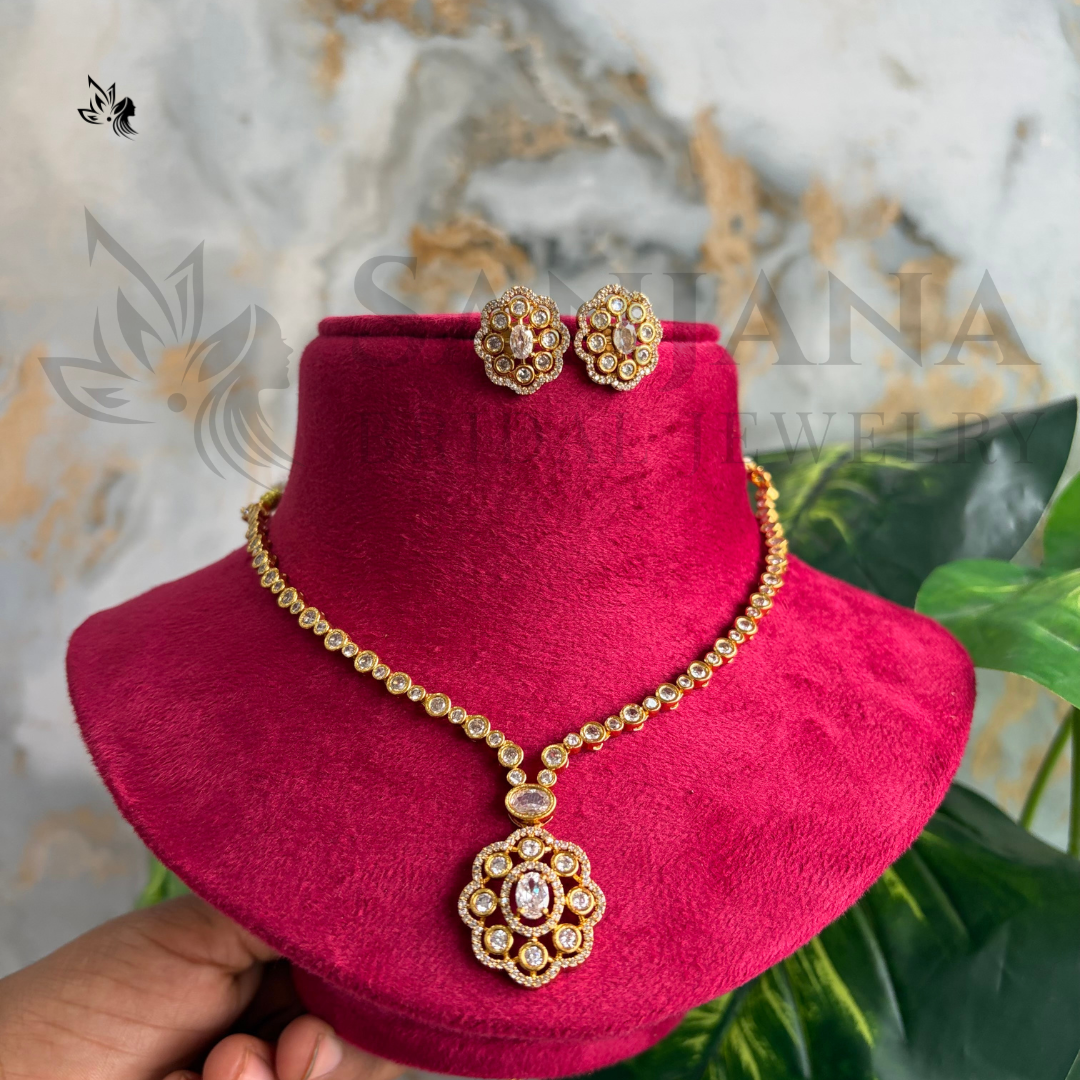Gold-toned jewelry set with a necklace and matching earrings displayed on a bright pink velvet stand. The jewelry features ornate floral motifs with clear stones and a central pink stone.