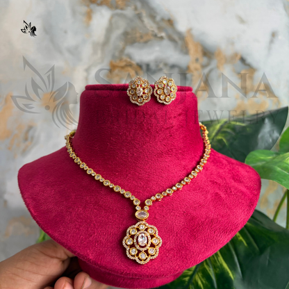 Gold-toned jewelry set with a necklace and matching earrings displayed on a bright pink velvet stand. The jewelry features ornate floral motifs with clear stones and a central pink stone.
