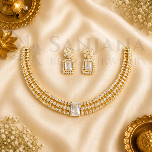 Gold jewelry set featuring a double-row necklace and matching drop earrings, embellished with clear stones, arranged on a beige satin fabric with decorative gold and white flowers.