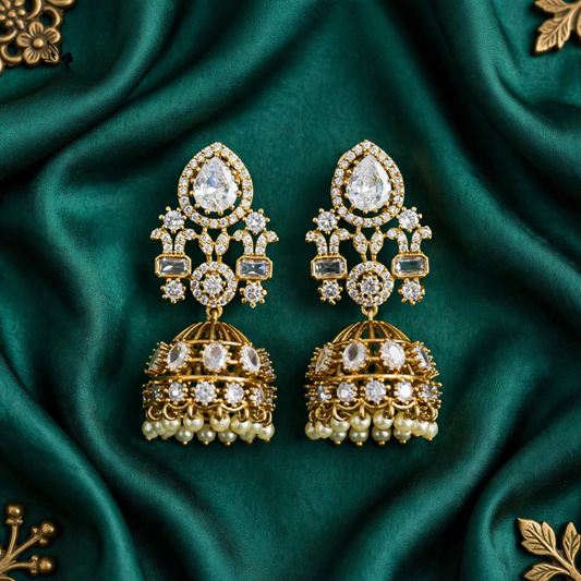 A pair of ornate gold-toned dangle earrings with clear gemstones and pearl-like drops, arranged symmetrically on draped emerald green satin fabric.