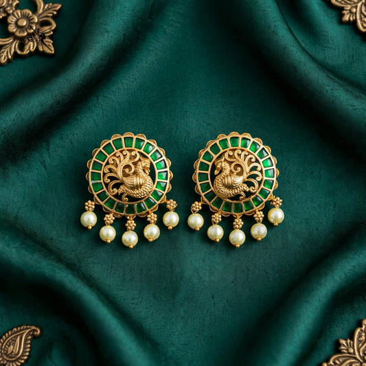 A pair of ornate gold-toned earrings with green enamel detailing, featuring a central peacock motif and dangling pearl-like beads, displayed on draped dark green fabric.