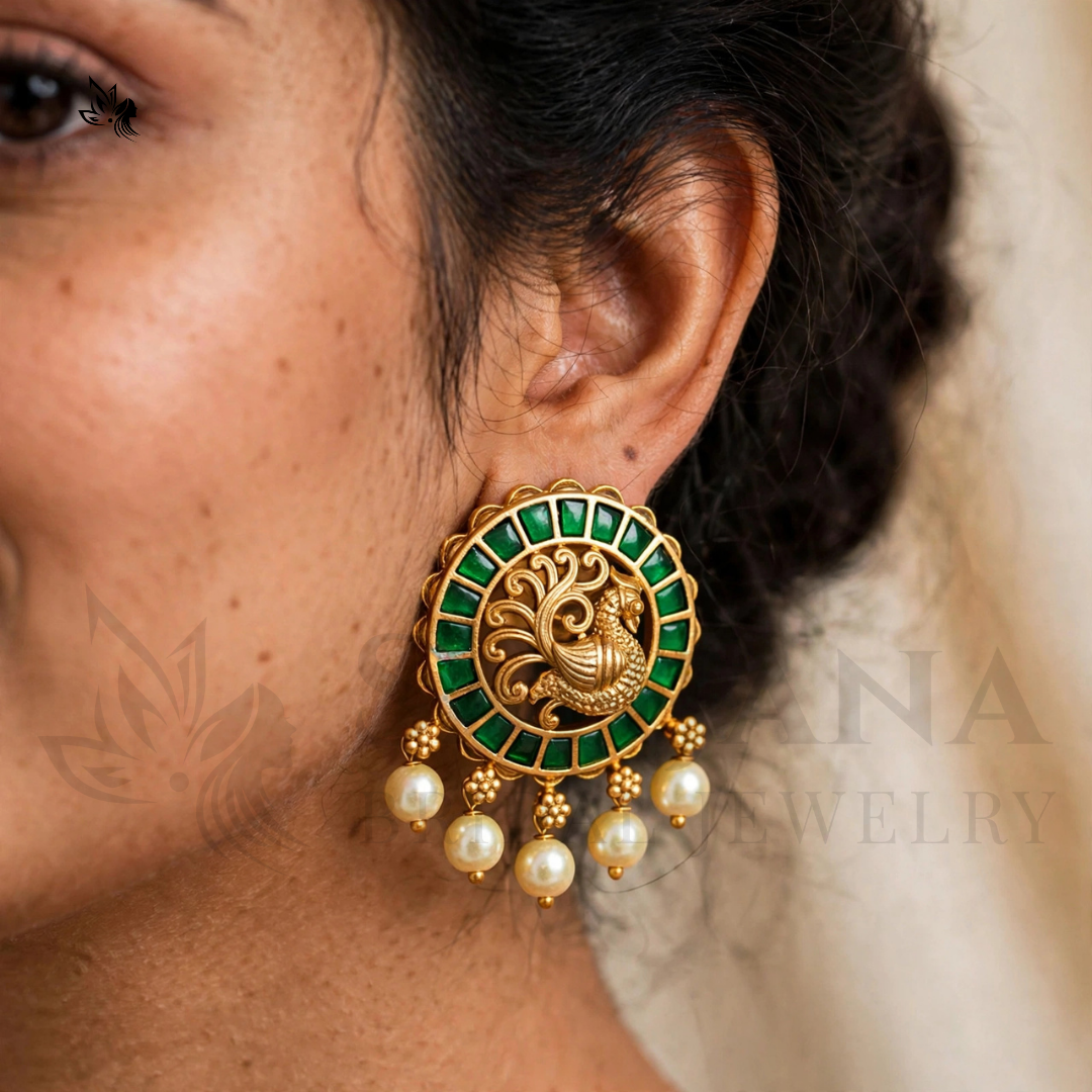 A close-up of a woman wearing a large gold-toned Indian-style earring with green square stones, a central floral motif, and three dangling pearl-like beads.