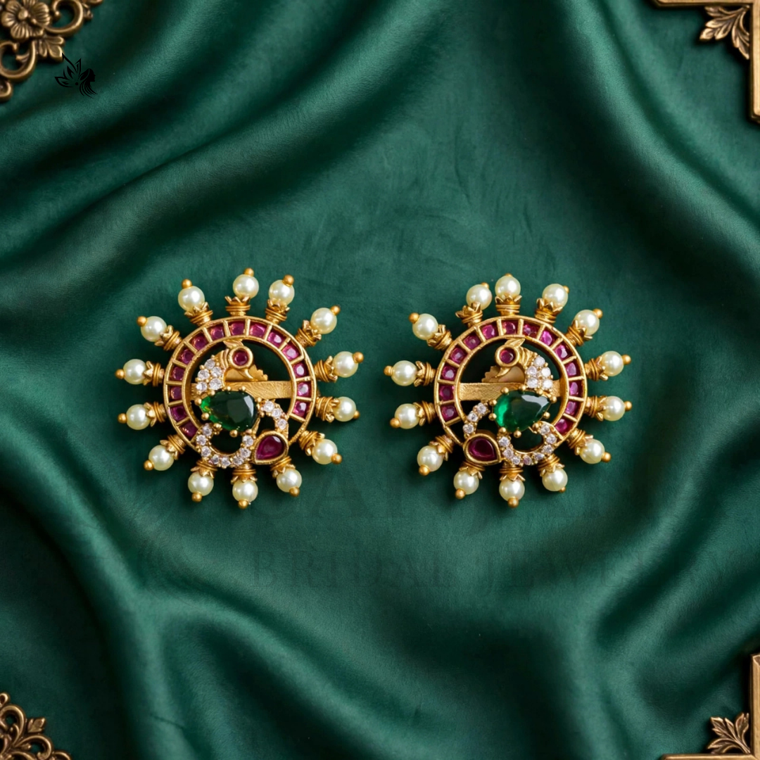A pair of ornate gold-toned earrings with red gemstone accents, green center stones, and white pearl-like beads, displayed on a draped dark green satin fabric.