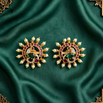 A pair of ornate gold-toned earrings with red gemstone accents, green center stones, and white pearl-like beads, displayed on a draped dark green satin fabric.