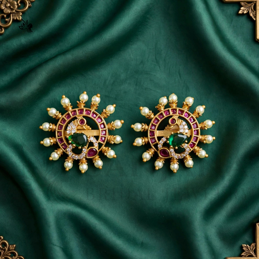 A pair of ornate gold-toned earrings with red gemstone accents, green center stones, and white pearl-like beads, displayed on a draped dark green satin fabric.