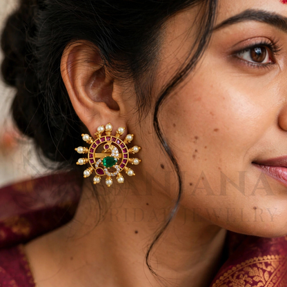 A close-up of a woman's ear wearing a large traditional Indian gold earring decorated with a green peacock motif, red stones, and white pearl-like accents.