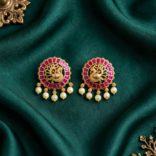 A pair of ornate gold-toned statement earrings with bright pink stone accents, a central deity motif, and dangling pearl-like beads, arranged on draped dark green fabric.