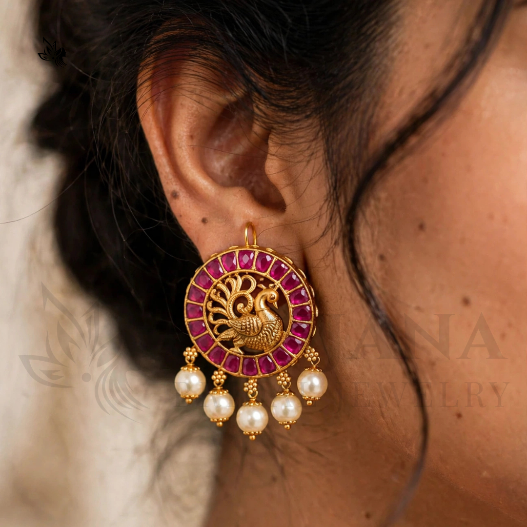 A close-up of a model's ear wearing a gold-toned dangle earring with a circular pavé pink stone design, a central peacock motif, and three dangling pearl beads.