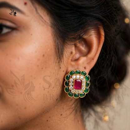 A close-up of a woman's ear wearing a gold-toned statement earring with a central rectangular pink gemstone surrounded by a halo of green stones and small clear stones.