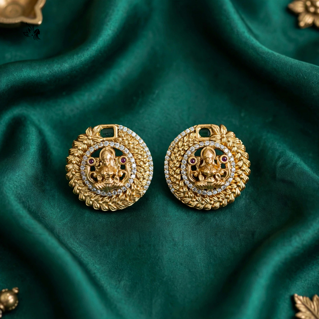 A pair of gold-toned traditional Indian earrings featuring a detailed Ganesha motif, set against a draped dark green satin fabric background.
