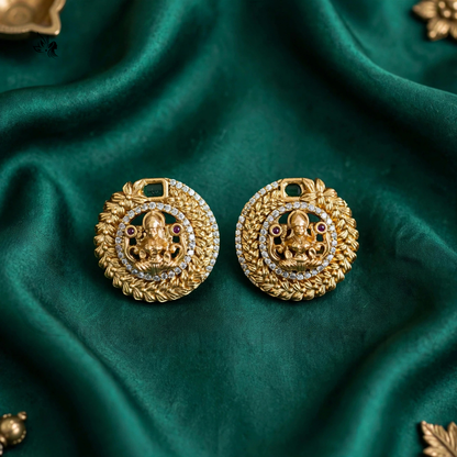 A pair of gold-toned traditional Indian earrings featuring a detailed Ganesha motif, set against a draped dark green satin fabric background.