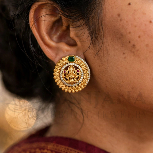 A close-up of a woman's ear wearing a detailed gold earring with a green stone and small red and white accents, against a softly blurred background.