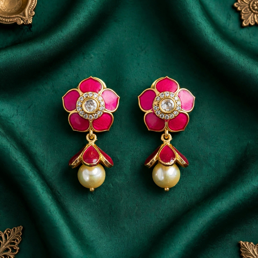 A pair of gold-plated floral dangle earrings with vibrant pink enamel petals, a central clear stone, and a dangling cream pearl, displayed on a dark green satin fabric background.