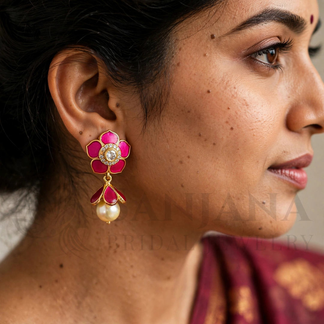 A close-up side profile of a woman wearing a gold-toned flower-shaped dangle earring with pink enamel petals, a central white stone, and a dangling pearl bead. The image has a faint watermark across it.