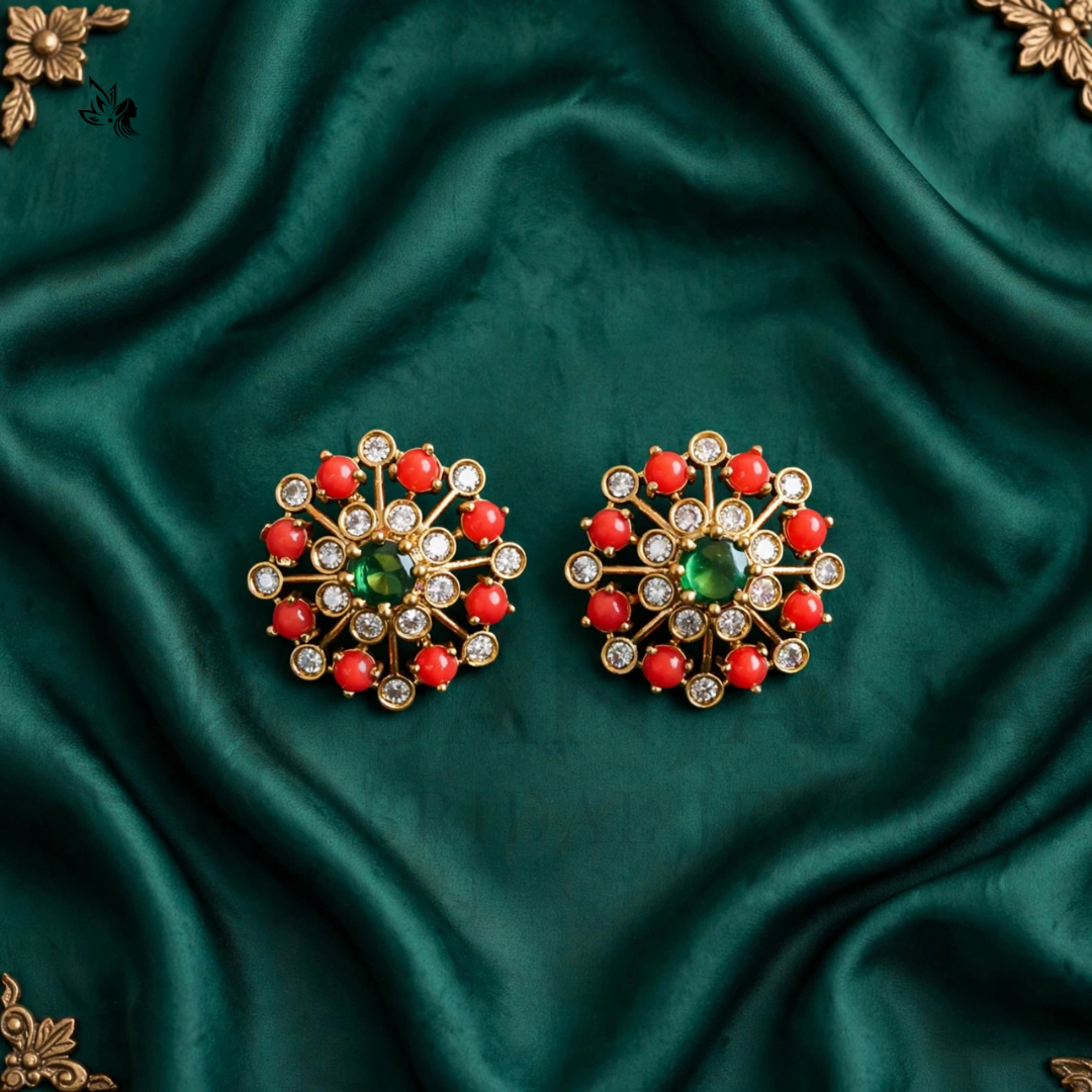A pair of ornate gold-toned statement earrings with a central green stone, surrounded by red stones and small clear stones, displayed on a draped dark green satin fabric.