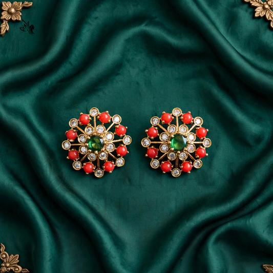 A pair of ornate gold-toned statement earrings with a central green stone, surrounded by red stones and small clear stones, displayed on a draped dark green satin fabric.