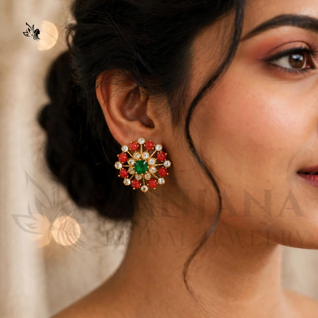 A close-up of a woman's ear wearing a decorative floral-shaped earring with a green center, red outer stones, and small white stones around a gold-tone setting.