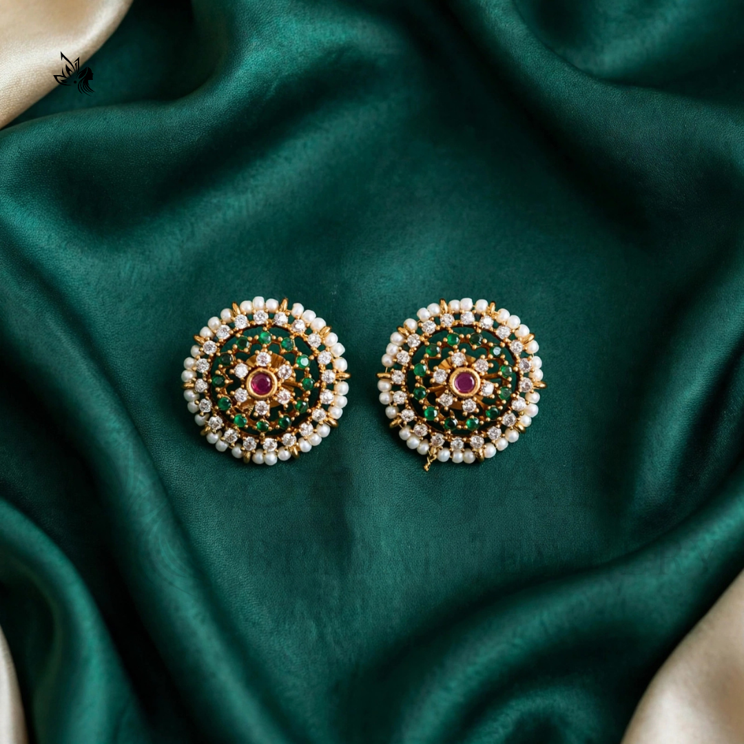A pair of ornate Indian Kundan earrings with green stone inlays, white pearl-like borders, and a central red stone on a dark green satin fabric background.