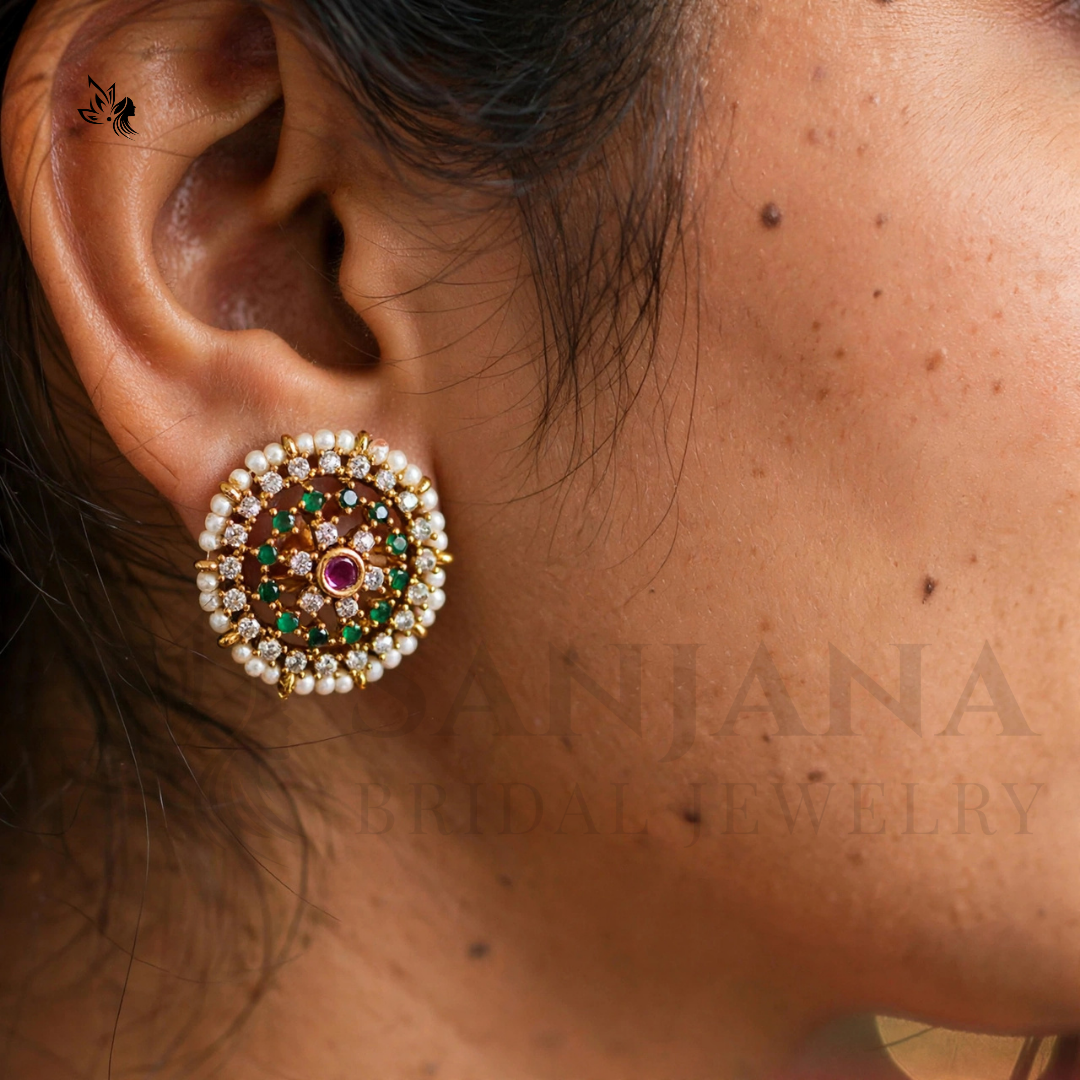 A close-up of a woman's ear wearing a round gold-toned earring adorned with white pearls, green stones, and a central red stone. A faint 'ANJANA' watermark is visible across the skin.