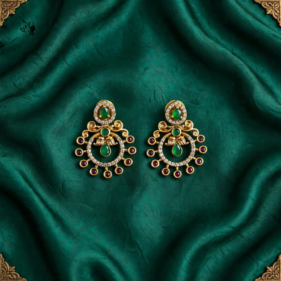 A pair of ornate gold-toned Indian chandelier earrings with green stones and small clear rhinestones, displayed on a draped dark green satin fabric.