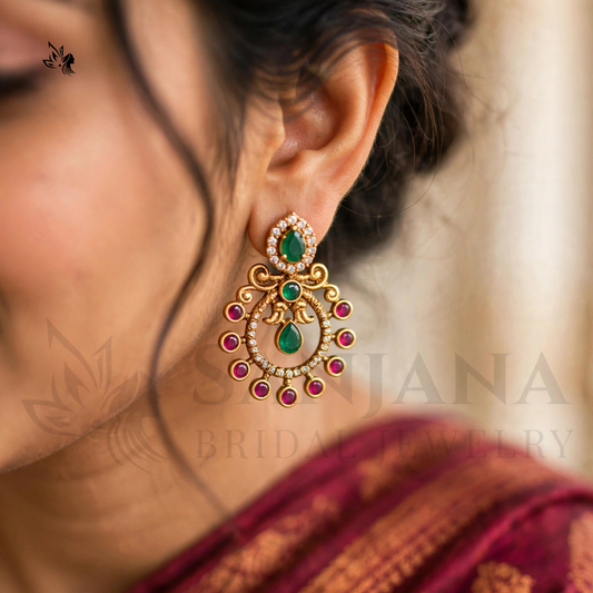 A close-up of a woman's ear wearing a large gold-toned traditional Indian chandelier earring with green and pink stones, set against a soft beige background.