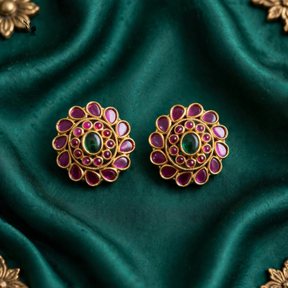 A pair of ornate gold-toned stud earrings with a central green oval stone and surrounding pink teardrop-shaped stones, displayed on a draped dark green satin fabric.