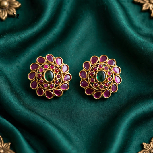 A pair of ornate gold-toned stud earrings with a central green oval stone and surrounding pink teardrop-shaped stones, displayed on a draped dark green satin fabric.