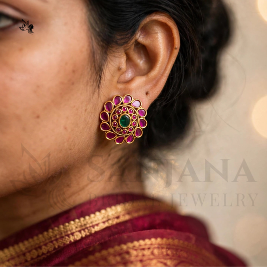 A close-up of a woman's ear wearing a traditional Indian gold earring adorned with pink stones, a green center stone, and gold filigree. The background is softly blurred with warm bokeh lights, and a faint 'SARAJANA JEWELRY' watermark is visible.