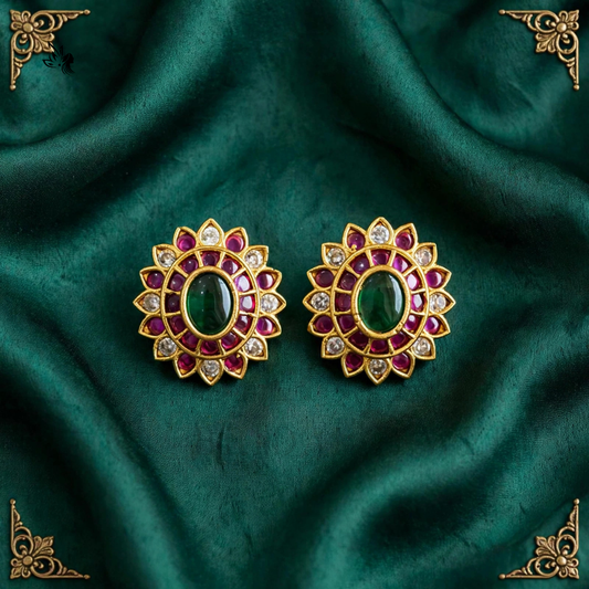 A pair of gold-toned Indian style earrings with oval green cabochon centers, surrounded by rows of red stones and clear accent stones, displayed on a draped dark green satin fabric.