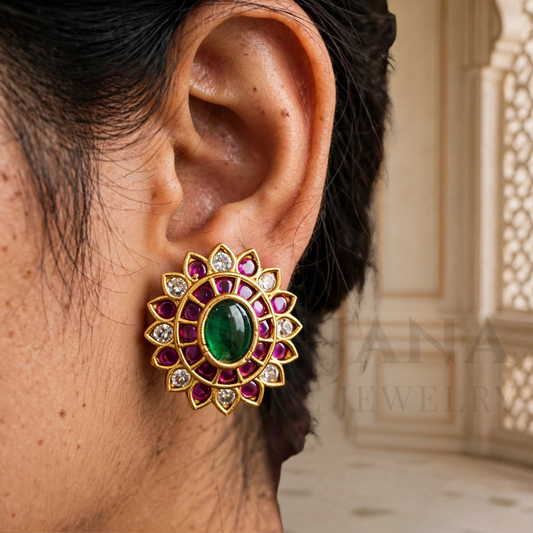 A close-up of a person's ear wearing a gold-toned floral stud earring with a central green oval stone, surrounded by pink gemstones and small clear stones, against an indoor architectural background.