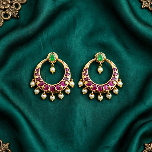A pair of gold crescent-shaped earrings with green teardrop stones, pink gemstone accents, and dangling white pearl beads, displayed on a draped dark green satin fabric.