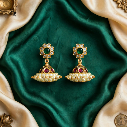 A pair of traditional Indian gold-plated Jhumka earrings with green and pink stones and white faux pearls, displayed on a dark green satin fabric with beige silk corner drapes.