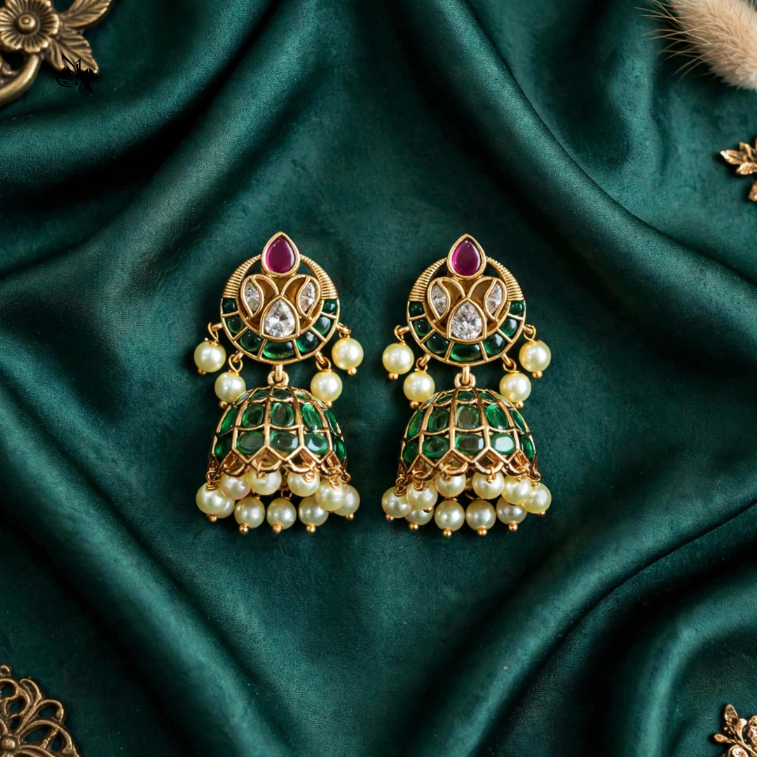 A pair of ornate gold-plated Indian jhumka earrings decorated with green enamel, white faux pearls, and a central pink stone, displayed on a draped dark green satin fabric.