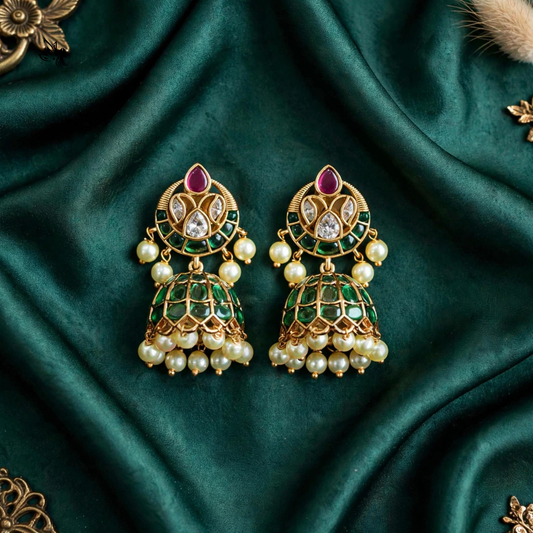 A pair of ornate gold-plated Indian jhumka earrings decorated with green enamel, white faux pearls, and a central pink stone, displayed on a draped dark green satin fabric.