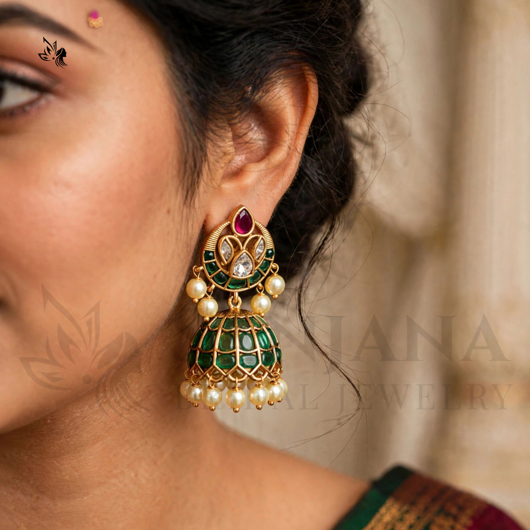 A close-up of a woman's ear wearing a gold-plated jhumka earring with green enamel, a red teardrop stone, and white faux pearl beads, against a soft beige background.