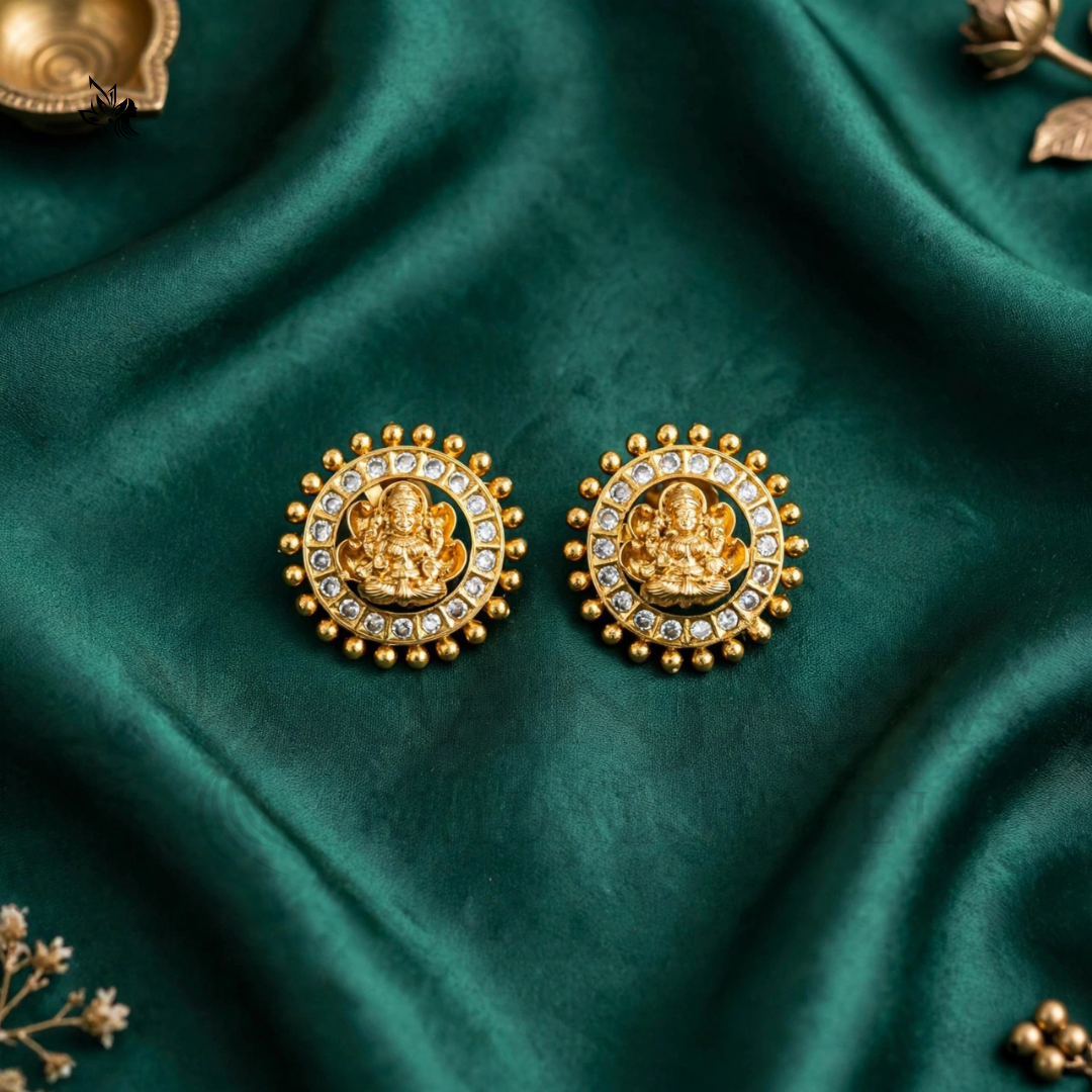 A pair of gold-toned circular earrings featuring a central deity motif surrounded by small clear stones and a beaded border, displayed on draped emerald green fabric.