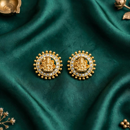A pair of gold-toned circular earrings featuring a central deity motif surrounded by small clear stones and a beaded border, displayed on draped emerald green fabric.