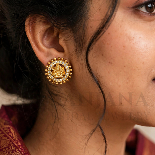 A close-up of a woman's ear wearing a gold-toned circular earring with a central deity motif and small clear stones around it. The image is cropped to show the ear, hair, and part of the face, with a faint watermark reading 'ANJUNA JEWELLERY' across the center.